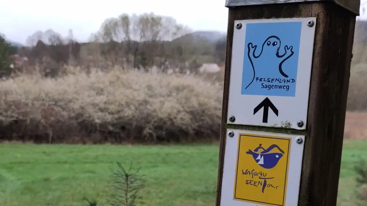 A small light blue metal sign on a wooden post with a blurry landscape in the background. The sign shows the stylized spirit marking the Felsenland Sagenweg. The name of the trail is written below the spirit.