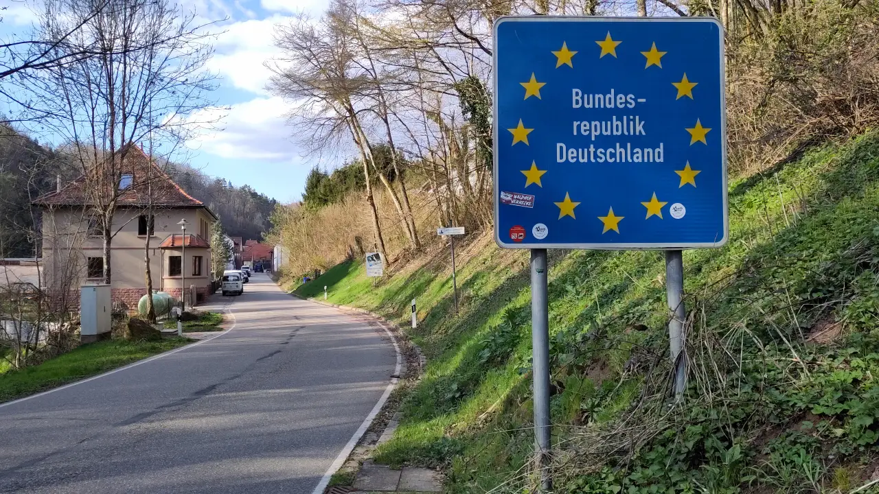 A street with a house in the background. In the foreground a blue sign with the circle of stars of the European Union and the text "Bundesrepublik Deutschland" ("Federal Republic of Germany")