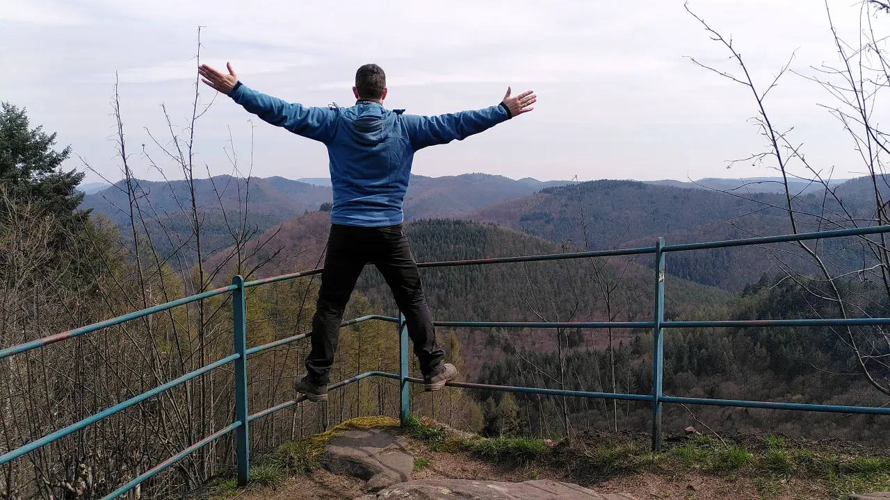 Markus from the back on the railing of the rock beneath the Peace Cross. You can see the hills of the Palatinate Forest.