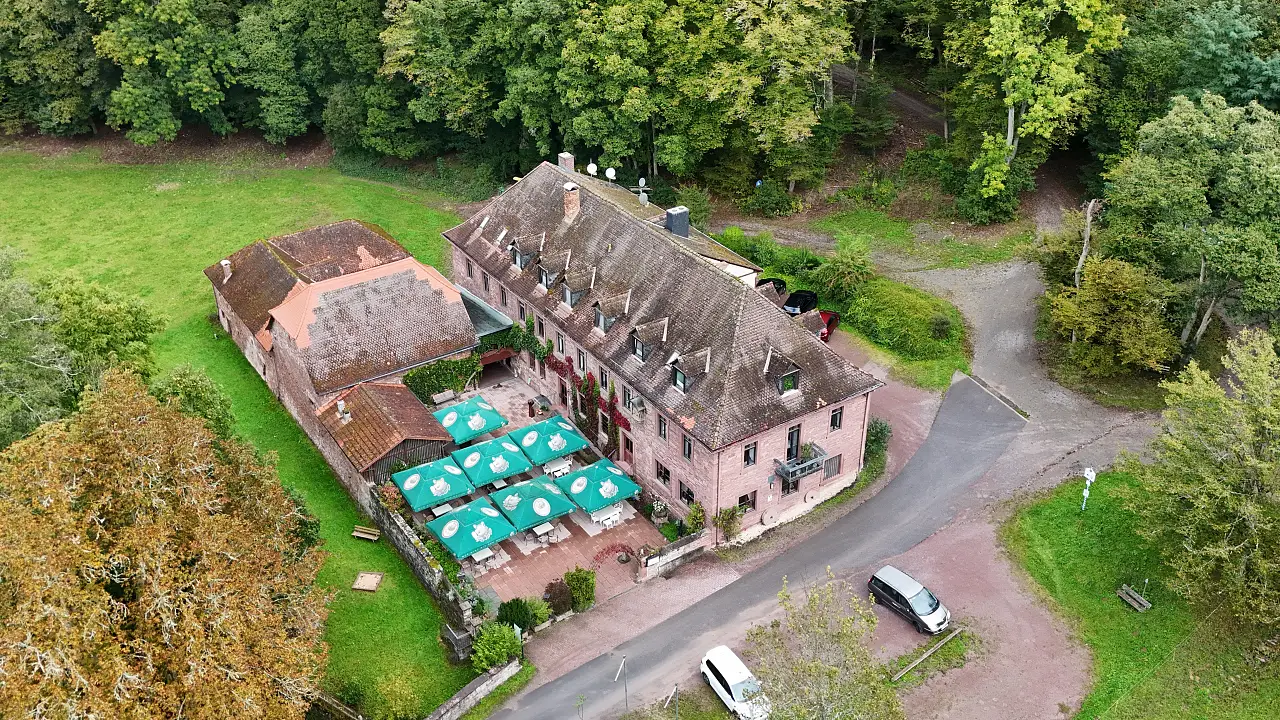 Der Gasthof Buchenmühle von oben. Ein altes Gebäude aus rötlichem Sandstein mit einem großen Nebengebäude und einigen großen Biergartensonnenschirmen dazwischen.