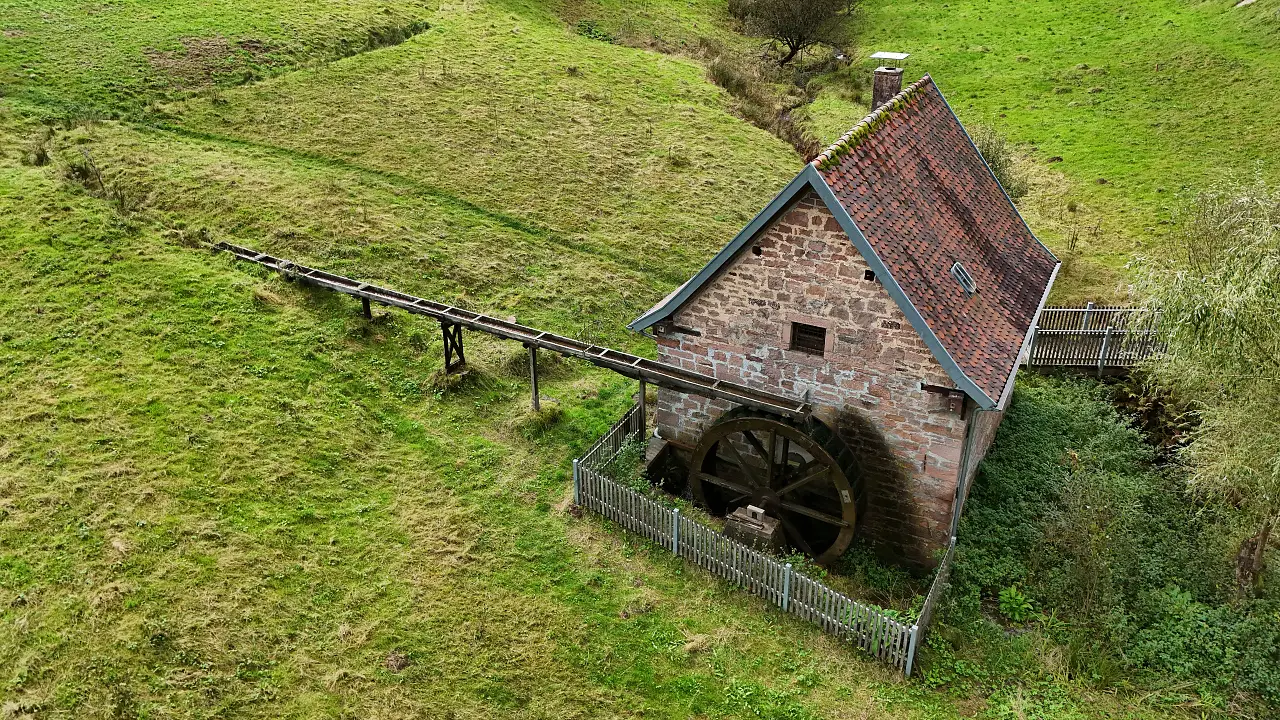 Die alte Dorfmühle in Habichtstal aus der Luft. Man sieht das alte Natursteingebäude mit dem Mühlrad an der Seite in der grünen Wiese.