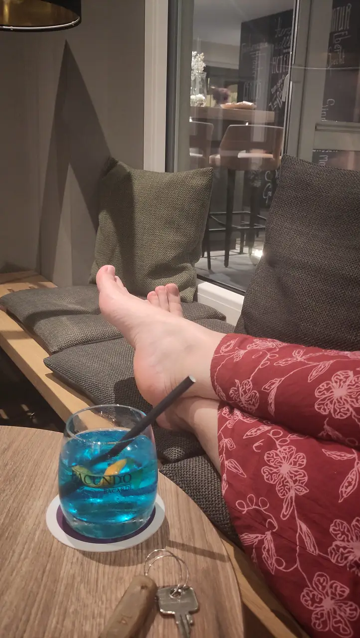 Übereinandergeschlagene Füße auf der Couch hochgelegt. Im Vordergrund ein Cocktail.