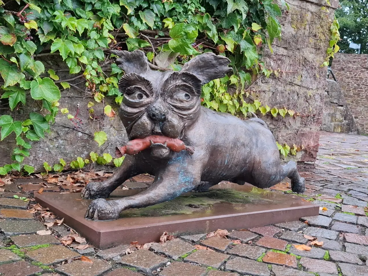 Eine Bronzestatue einen kleinen Hundes, der mit einer Wurst im Maul davonrennt