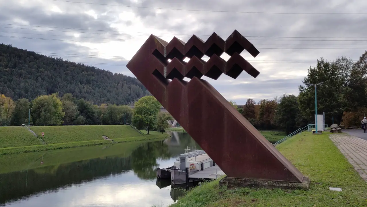 Eine Skulptur aus verrostetem Stahl. Eine schräg aufragende Stahlsäule mit stilisierten, eckigen Wellen an der Spitze. Im Hintergrund das Wasser und die Deiche des Schutzhafens in Gemünden