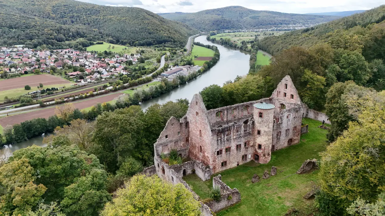 Blick auf die Ruine Schönrain am Main. Ein dreistöckiges Gebäude mit hohen Giebeln ohne Dach. An der einen Wand ein runder Turm mit einem Kupferdach. Das gesamte Gelände ist mit Gras bewachsen und von Bäumen umgeben. Im Hintergrund schlängelt sich der Main durch's Tal